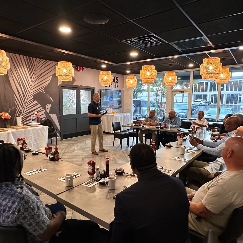 A man presents to a group of men seated at tables in Mom's Kitchen, a breakfast restaurant.