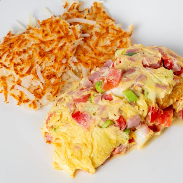 A Mom's Kitchen breakfast plate with a fluffy omelet filled with ham, tomatoes, and green onions, alongside crispy hash browns.