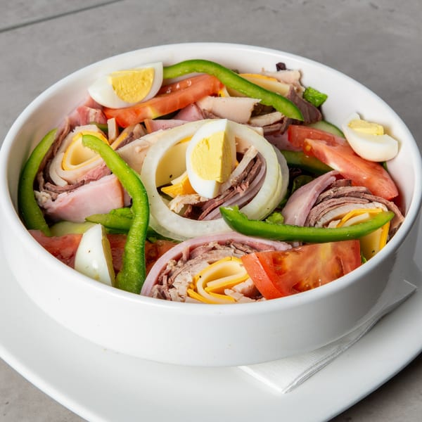 A large salad bowl filled with sliced meats, hard-boiled eggs, tomatoes, onions, and green peppers.
