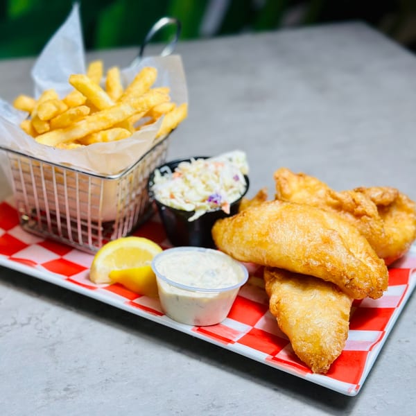 Golden-brown fish and chips with coleslaw, tartar sauce, and lemon on a red checkered plate.