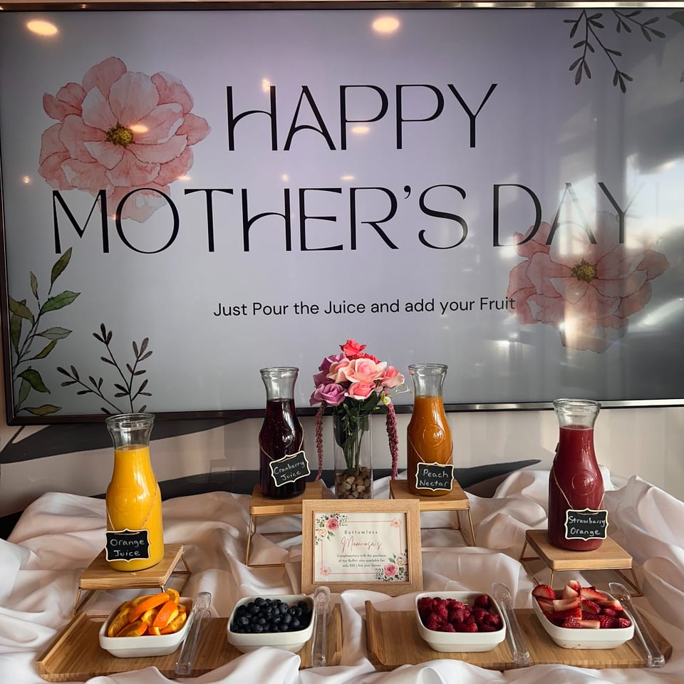 A Mother's Day mimosa bar with various juices, fresh fruit, and a sign for "Bottomless Momosas".
