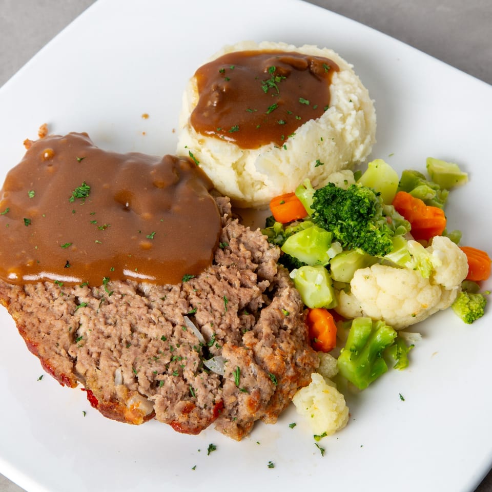 Meatloaf with gravy, mashed potatoes, and mixed vegetables on a white plate.