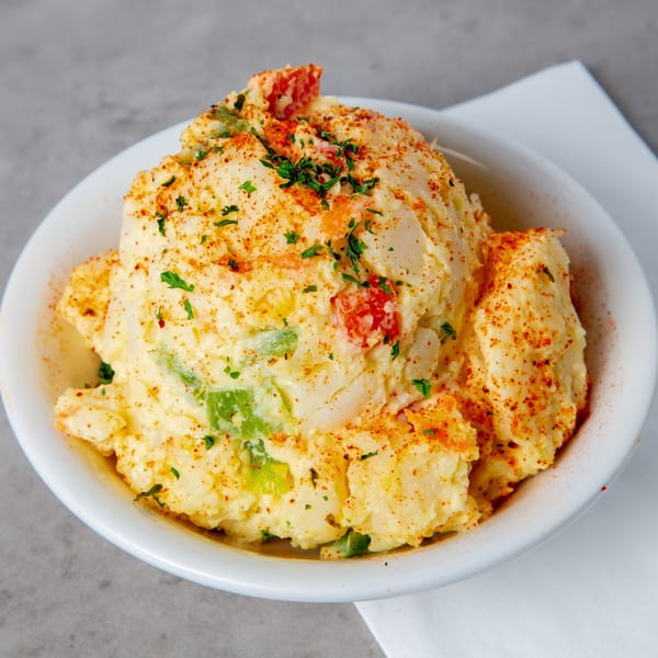 Creamy potato salad with red and green peppers, sprinkled with paprika and parsley in a white bowl.