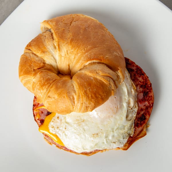 A Mom's Kitchen breakfast croissant sandwich with ham, fried egg, and melted cheese on a white plate.