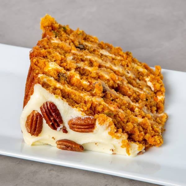 A slice of Mom's Kitchen carrot cake with cream cheese frosting and pecans on a white plate.